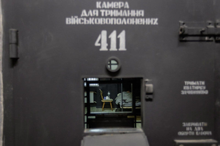 A view through a small hatch in a heavy metal door marked ‘411’ shows a sparse prison cell with a chair and a cot where a person is lying under a blanket