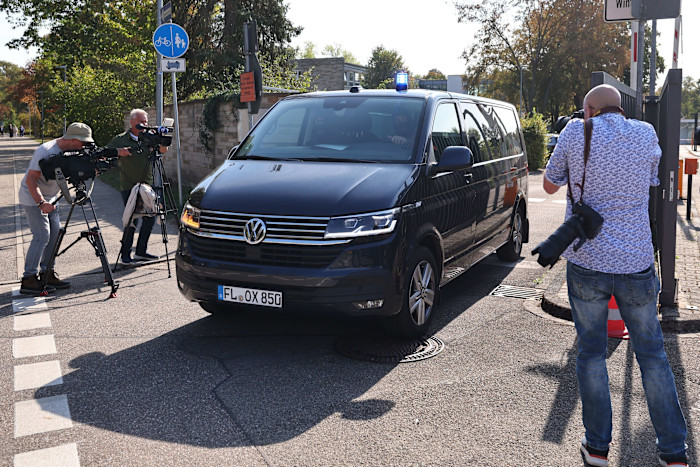 A black Volkswagen van with a blue light on top leaves the court, surrounded by photographers and videographers capturing the scene.