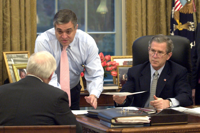 President George W. Bush sits at his desk as George Tenet speaks to Dick Cheney, who has his back to the camera.