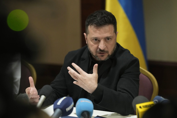 Volodymyr Zelenskyy gestures while speaking at a news conference, seated at a table with microphones in front of him.
