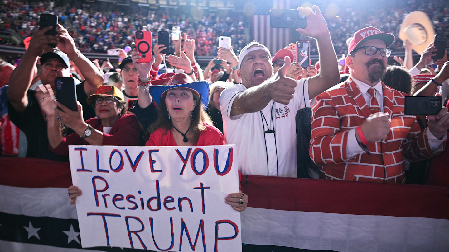 Supporters at a rally cheer, hold up phones, and display a handmade sign reading “I LOVE YOU President TRUMP” as Donald Trump arrives to speak.