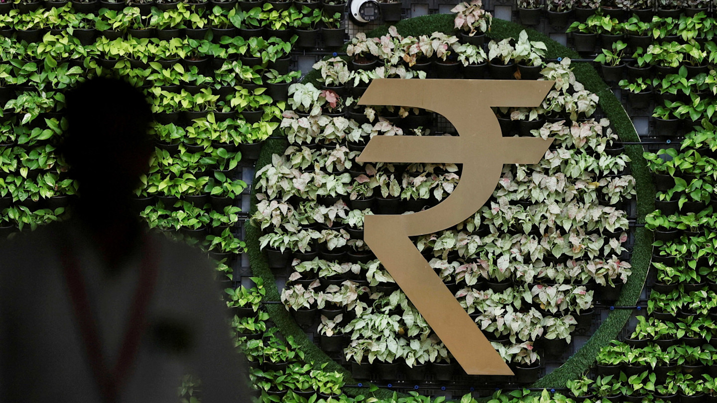 A large gold Indian Rupee symbol displayed on a wall of green plants, with a silhouetted man walking past in the foreground.