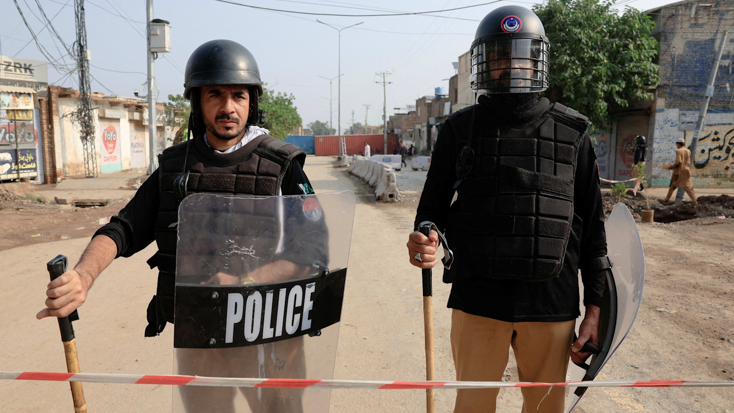 Two police officers in riot gear stand behind a barricade, blocking a road with shipping containers in the background.
