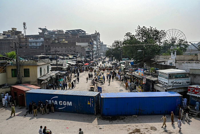 Shipping containers block a road as people walk around and gather amid shops during protests in Peshawar. Police officers stand near the containers.