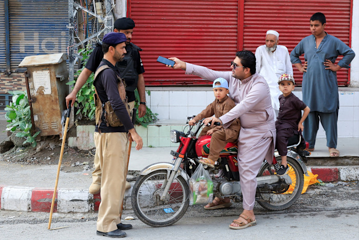 A man on a motorcycle with two children shows a phone to police officers guarding a road, while bystanders watch nearby.