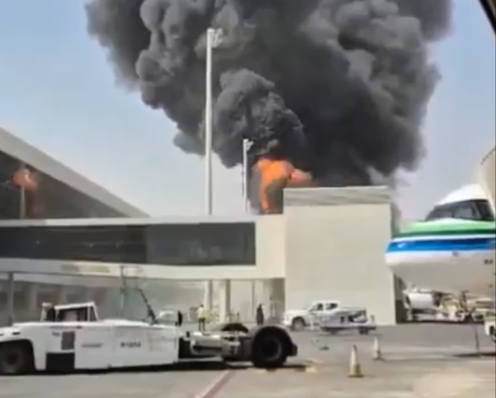 Thick black smoke and flames rise from a building at King Khalid International Airport, with airport vehicles nearby.