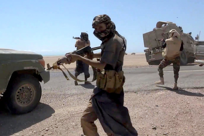Armed fighters in military gear take cover and fire weapons near vehicles on a desert road during clashes in Yemen.