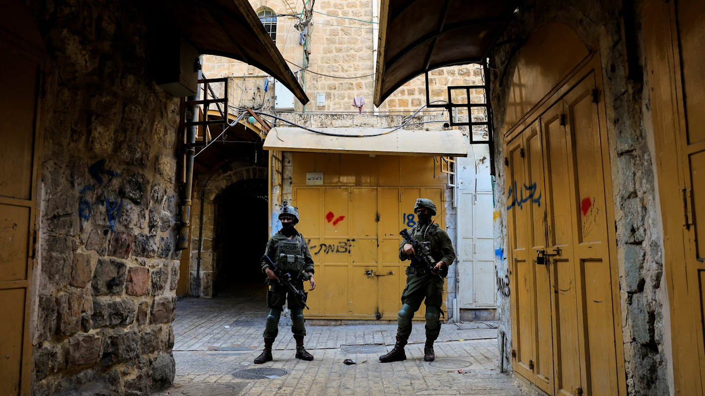 Two Israeli soldiers in uniform and protective gear stand guard in an alley with closed yellow metal shop doors in Hebron.