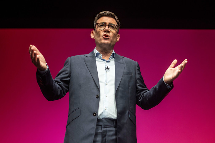 Andy Burnham delivers a keynote speech with arms raised, standing in front of a solid pink background.