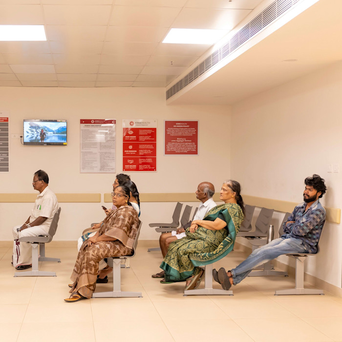 Several people sit spaced apart in a hospital waiting area, some looking forward and others looking at their phones.