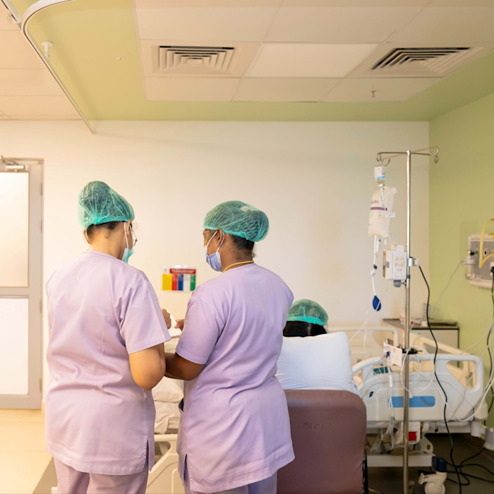 Two nurses in scrubs and hair covers consult near a patient’s bedside in a hospital room.