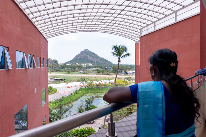 A woman in a blue sari stands at a railing inside KIMS Health Nagercoil, looking out towards a green hill and palm tree.