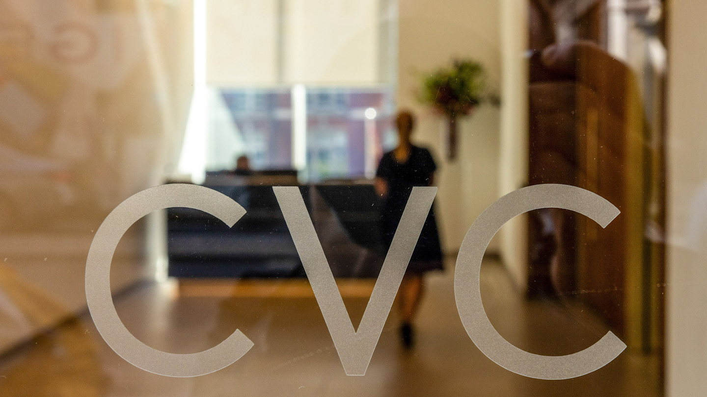 CVC Capital Partners logo seen on glass with a blurred office lobby and two people in the background.
