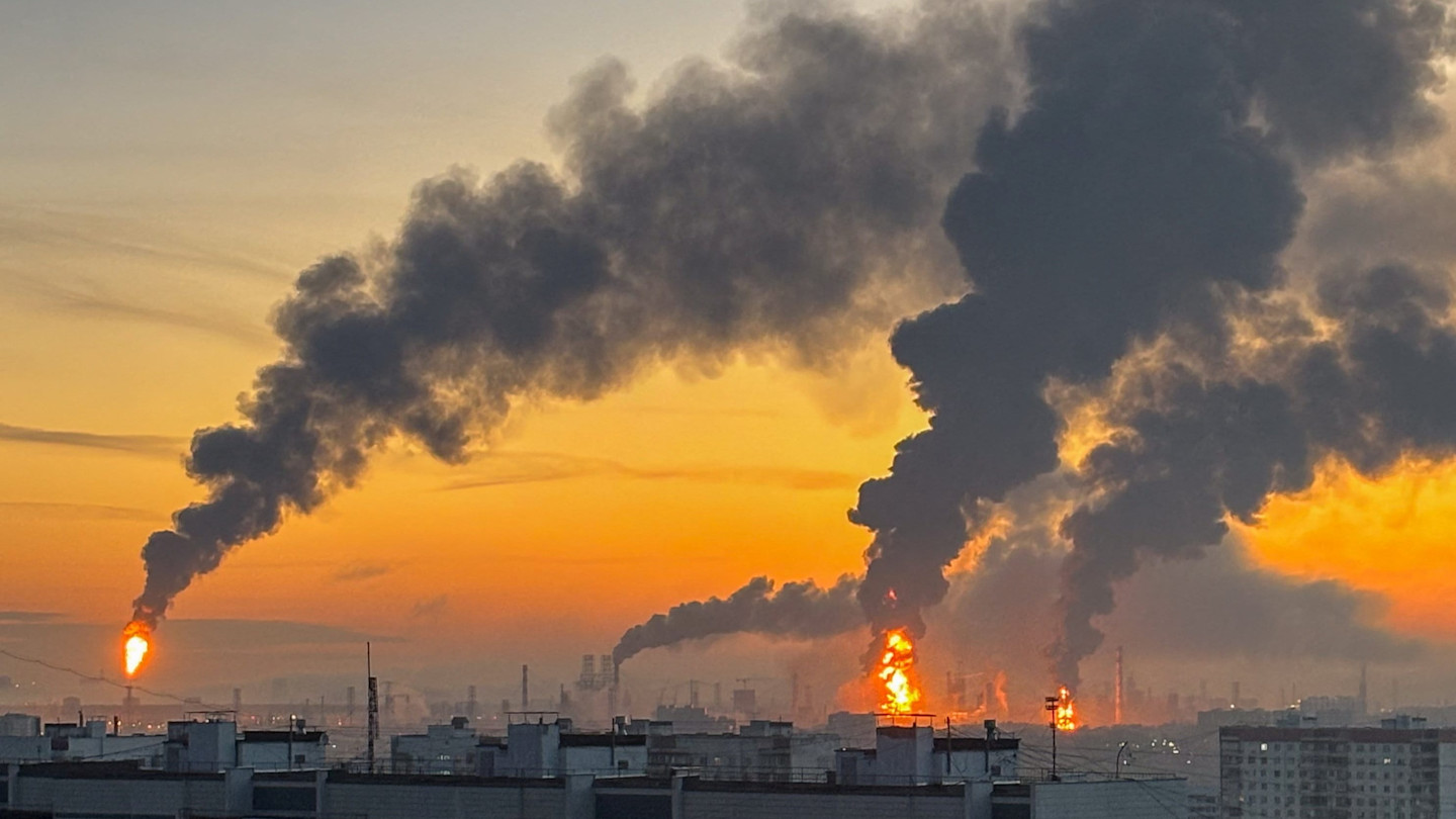 Plumes of smoke and flames rise from multiple fires at an oil refinery against an orange sunset sky.