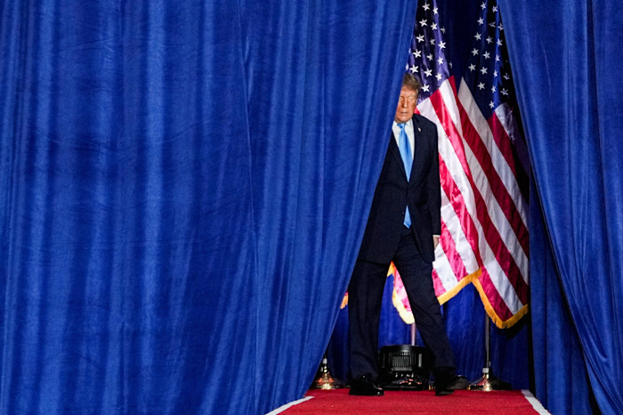 Donald Trump walks onstage between blue curtains, with American flags visible behind him.