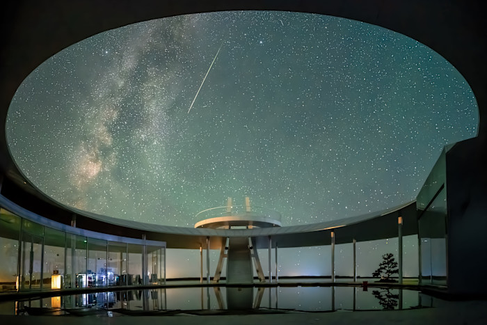 A bright meteor streaks across a star-filled night sky above a modern, circular lakeside pavilion with glass walls.