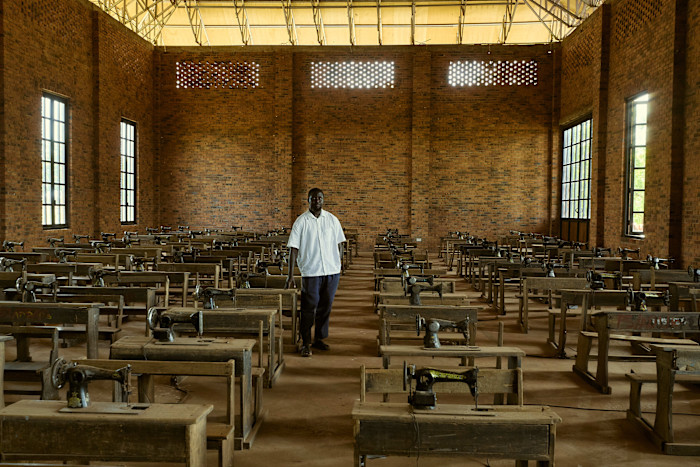 Ibrahim Mahama stands amid an art installation comprising rows of old school desks each fitted with a sewing machine