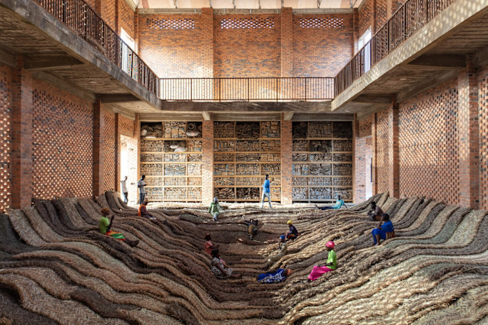 People sit and walk on undulating woven hay mats inside a large brick hall, with shelves of found objects lining the back wall.