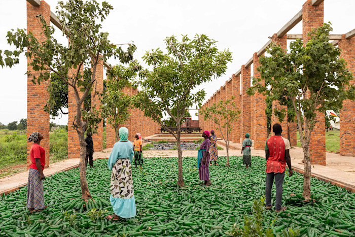 Several people stand among trees on a ground covered with green bottles, surrounded by tall brick pillars in an outdoor installation.