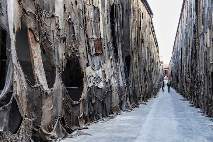 Huge installation by Ibrahim Mahama made of stitched old sacks covering the walls of a long alleyway, with two people walking in the distance.
