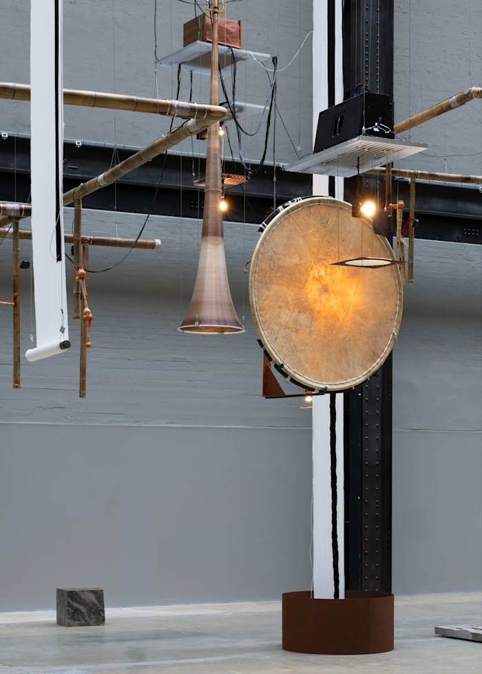 A large drum, wooden horn, and bamboo pipes are suspended in an industrial gallery space as part of Tarek Atoui’s installation.