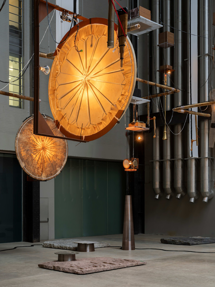Large circular drum-like installations hang from a metal framework, illuminated from within, surrounded by pipes and scattered stone slabs on the floor.