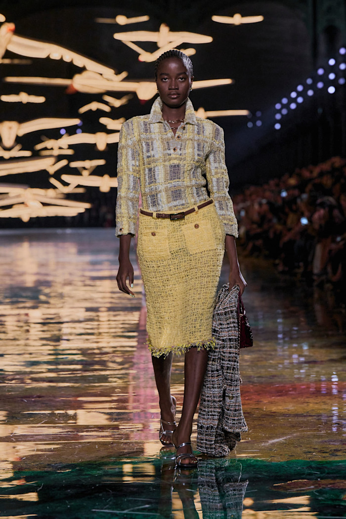Model Rachel Ojuromi on the catwalk in a grey and yellow checked top and yellow skirt, holding a black and white checked jacket and black handbag.