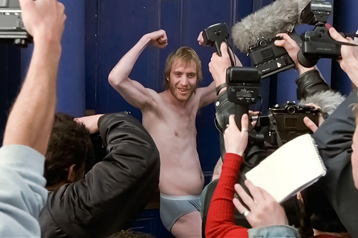 Rhys Ifans poses in underwear and flexes his arms while surrounded by photographers and reporters in a scene from "Notting Hill."