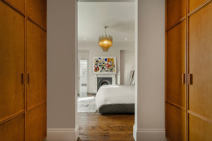 A view through a hallway with wooden wardrobes into a bedroom featuring a chandelier, a modern bed and a colourful abstract painting above a fireplace.
