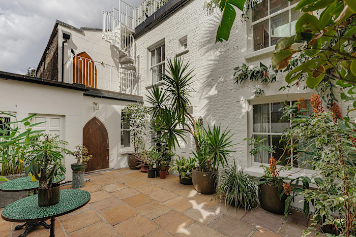 A private courtyard with potted tropical plants, stone paving, white-painted brick walls, and a white spiral staircase.