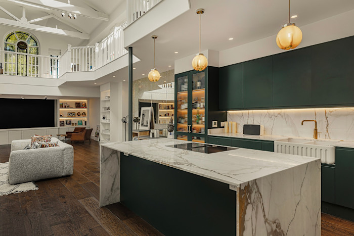 Modern open-plan kitchen and living area with high ceilings, marble countertops, dark green cabinets and gold light fixtures.