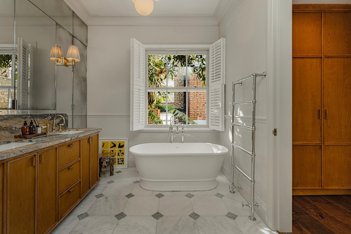 A bright bathroom with a freestanding bathtub beneath a window with open shutters, marble flooring, wooden cabinets and a chrome towel rack.