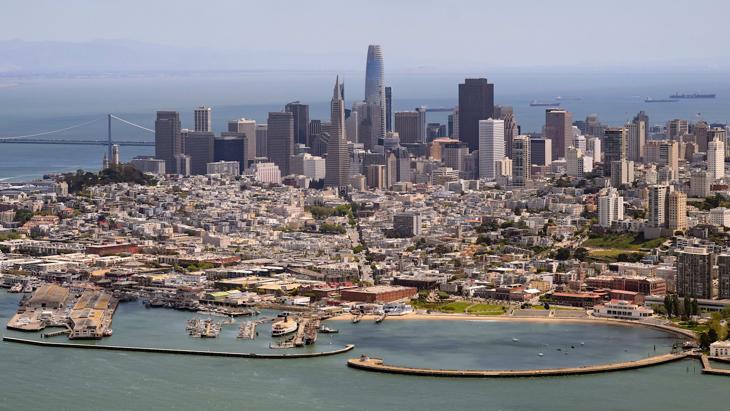 Aerial view of downtown San Francisco with the city skyline, waterfront, and surrounding bay visible under clear skies.