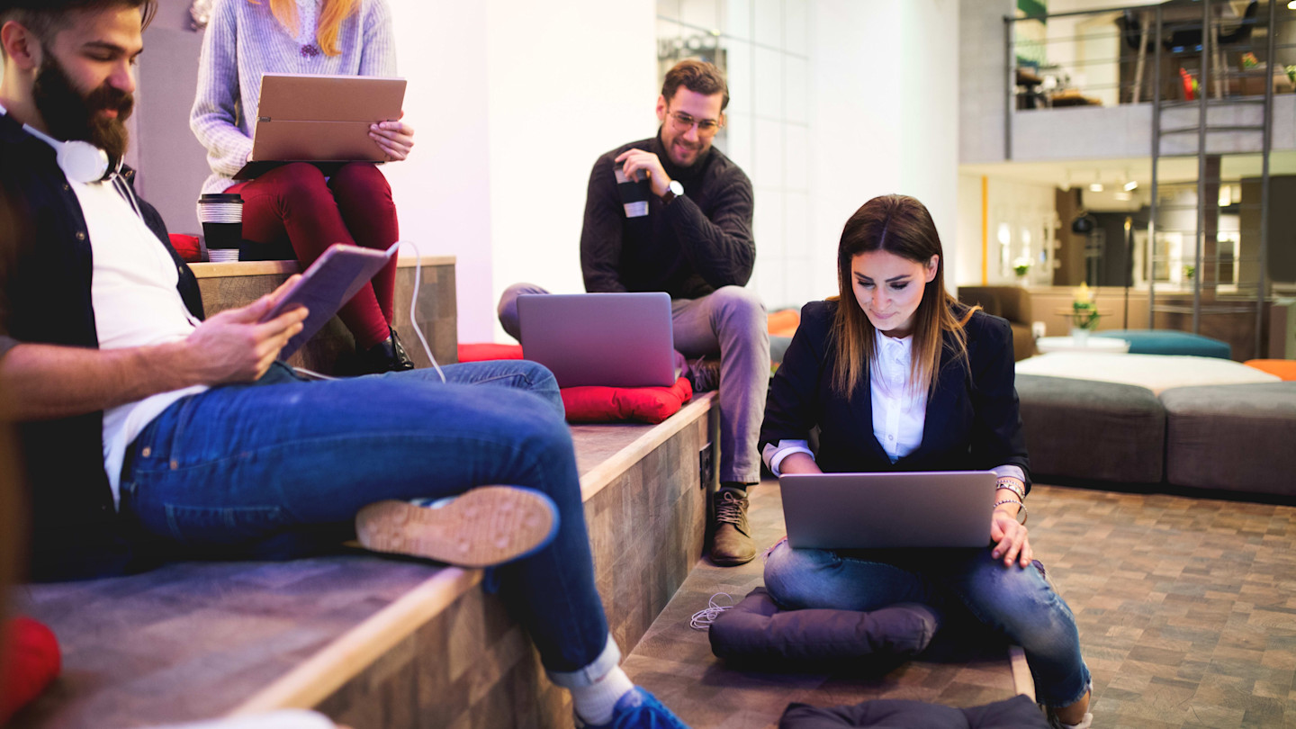 Four people working together with laptops and tablets in a modern, informal workspace.