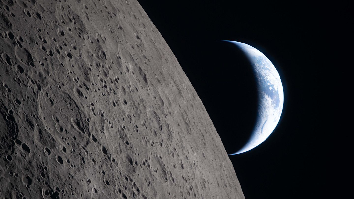 A crescent Earth is visible above the Moon’s cratered surface, with the Ohm crater in the foreground.