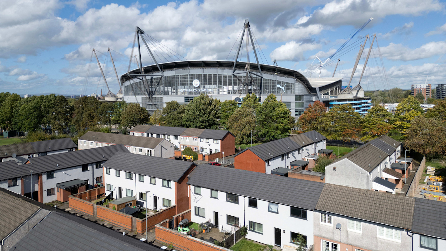 Aerial view of Manchester City’s Etihad Stadium with rows of residential houses and trees in the foreground.