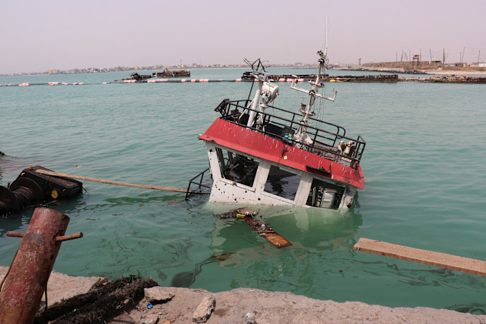The navigation bridge of a sunken vessel is partially submerged and tilted in the water near a dock, with debris floating nearby.