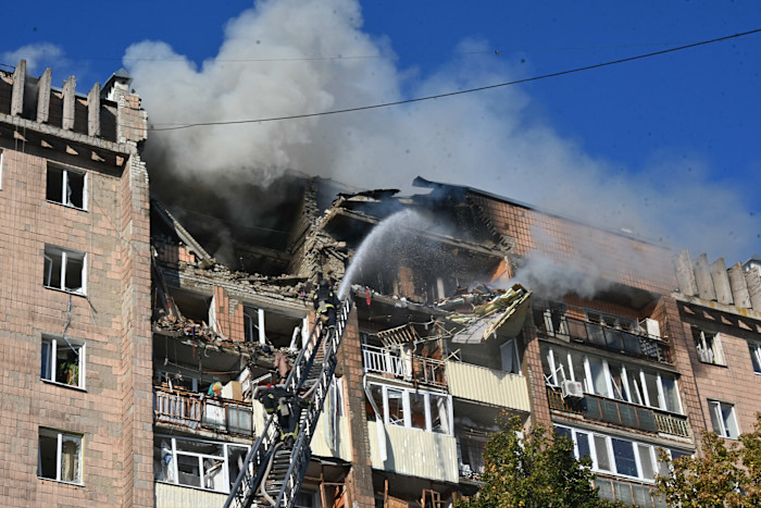 Firefighters on a ladder spray water into the upper floors of a residential building heavily damaged and smoking after a drone strike.
