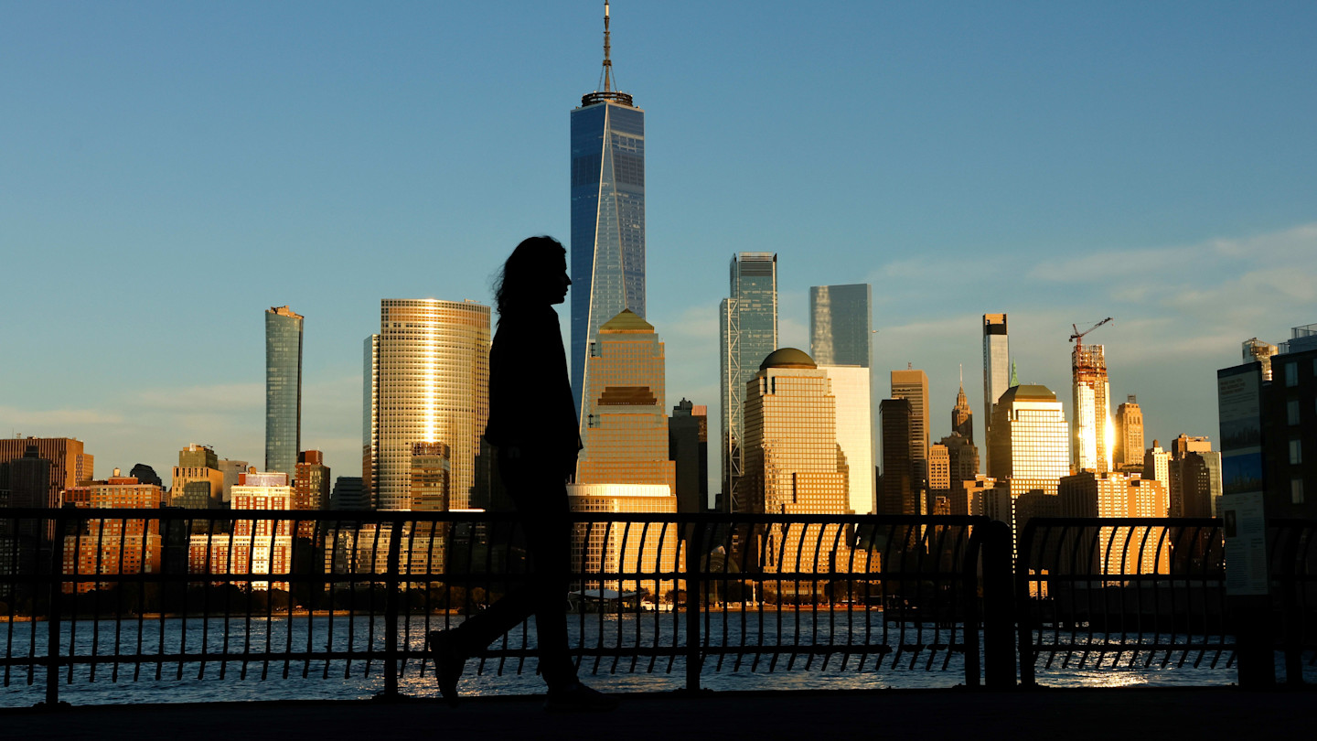 A person is silhouetted while walking along the Hudson River with the lower Manhattan skyline and One World Trade Center at sunset.
