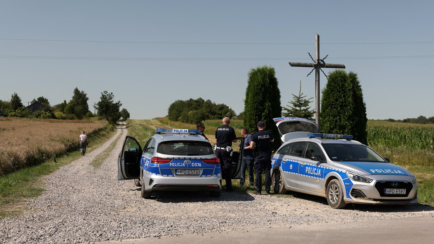 Several police officers stand near two police vehicles parked on a rural road by a field, next to a large cross and trees.
