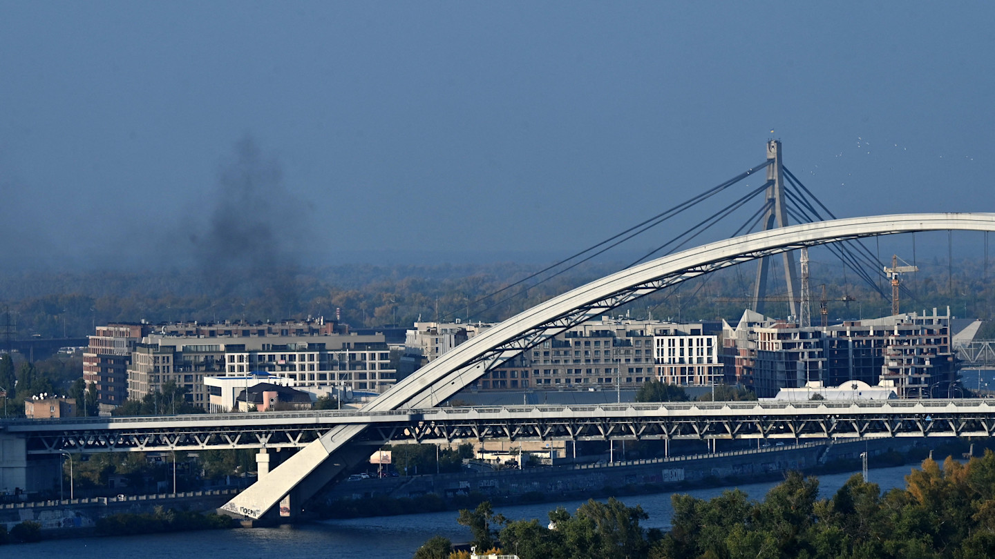 Black smoke rises above buildings in Kyiv after a drone explosion, with a large bridge and river in the foreground.