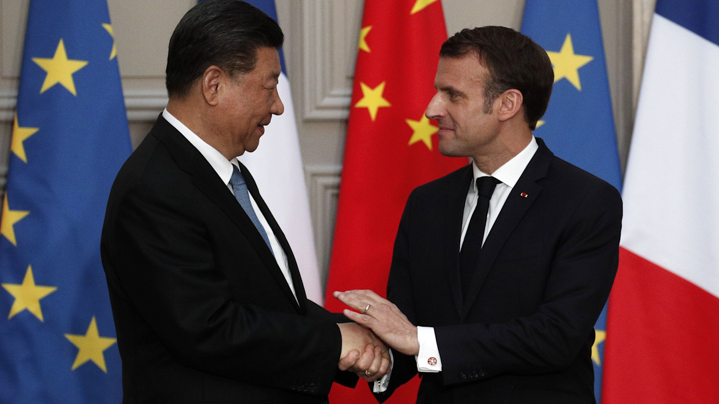Emmanuel Macron and Xi Jinping shake hands and smile at each other in front of French, Chinese and EU flags