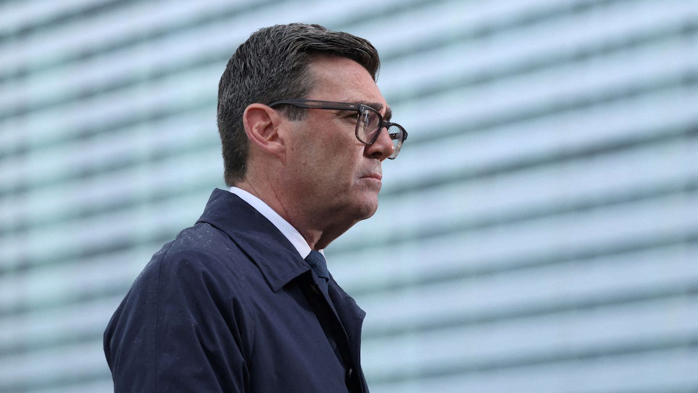 Andy Burnham stands in profile, wearing glasses and a dark coat, following a meeting.
