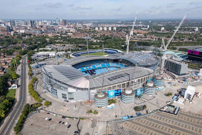 Aerial view of Etihad Stadium in Manchester, showing the football pitch, stands and ongoing construction with cranes around the venue.