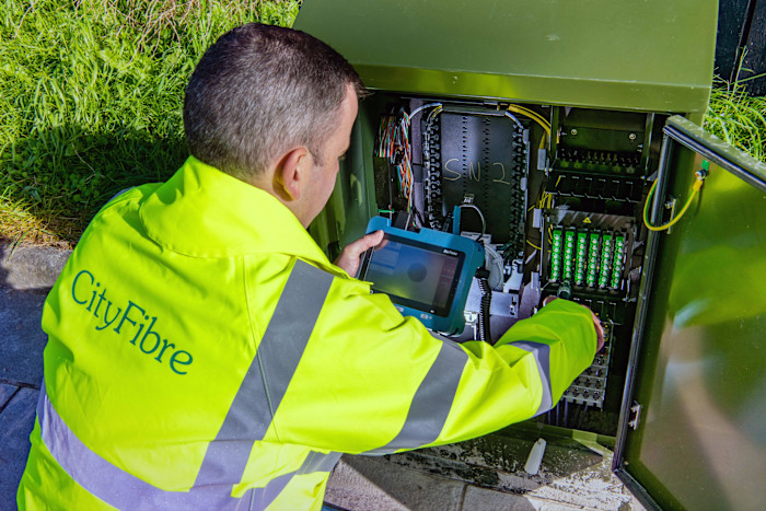 A person in a CityFibre jacket works with equipment inside an open street cabinet at the Raploch estate installation site.