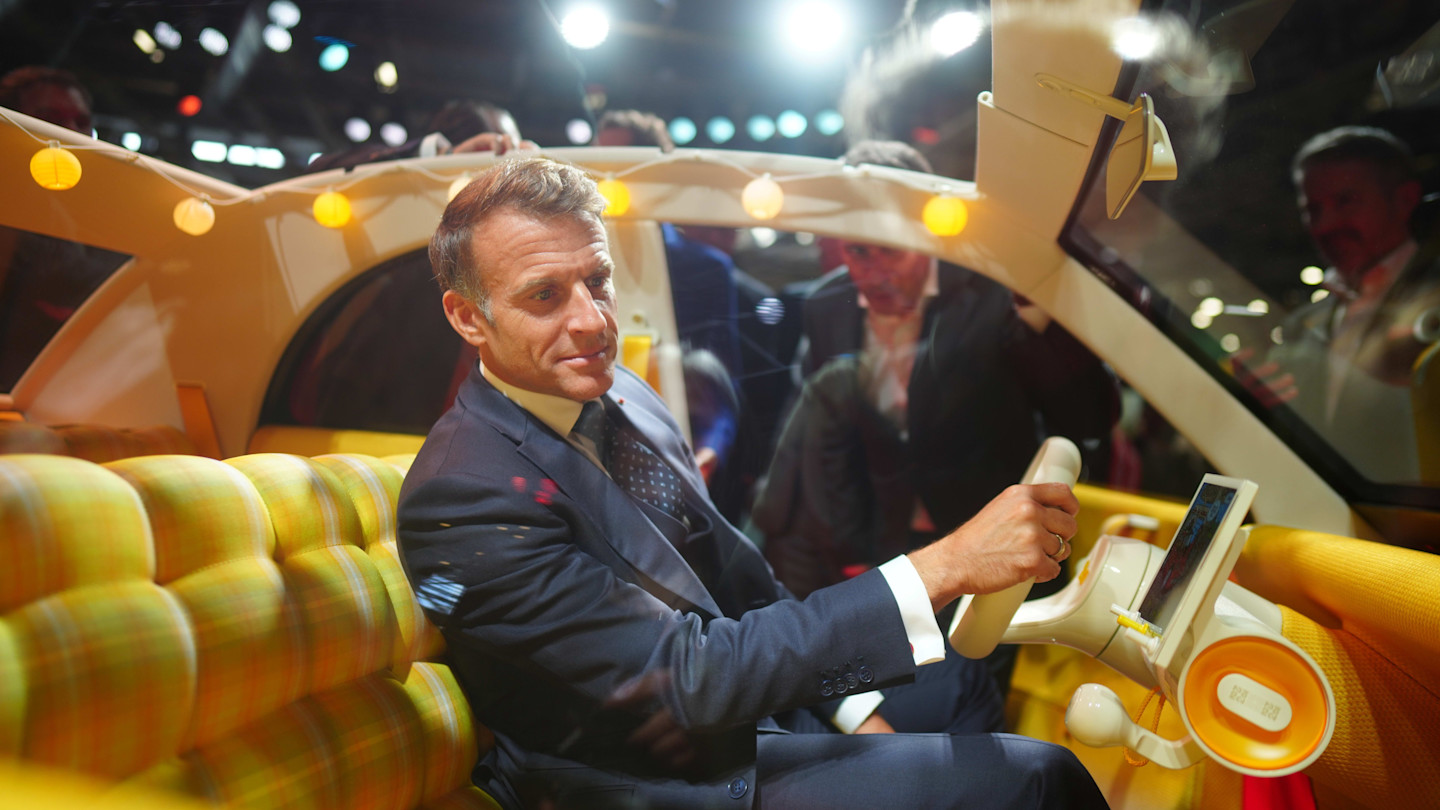 Emmanuel Macron sits in the driver’s seat of a Citroën 2CV, holding the steering wheel at the Paris Motor Show.