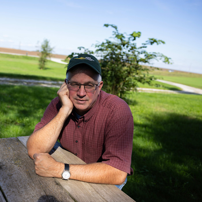 Aaron Lehman in collared shirt and peaked cap sits at an outdoor table on his farm