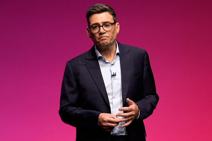 Andy Burnham delivers a keynote speech against a pink background, wearing a dark suit and glasses, with a microphone clipped to his shirt.