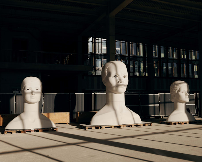 Three large white busts of women with stylised features are displayed on wooden pallets inside a studio with industrial lighting.