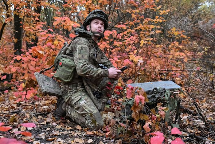 A Ukrainian artilleryman in camouflage uniform kneels among autumn foliage, setting up a Starlink satellite dish near the frontline.